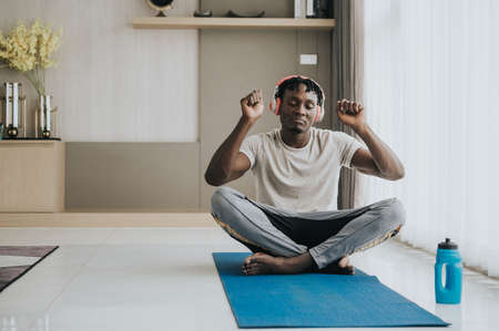 African man wearing sport outfit dancing while using headphone to listen music exercise yoga workout at home for good healthの写真素材