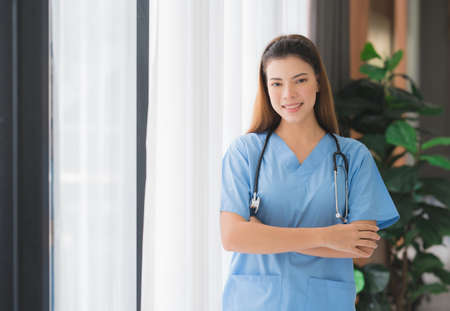 Portrait of happy smiling Asian beautiful nurse or caregiver woman standing  by the window crossed arms with stethoscopeの写真素材