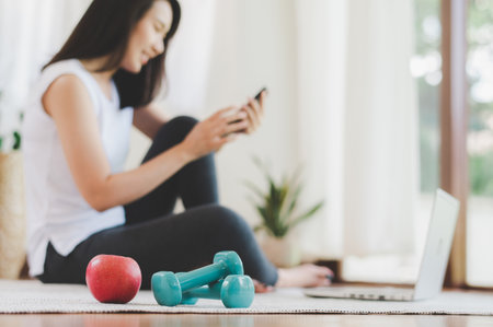 apple and dumbbells with happy healthy asian woman using smartphone and laptop after fininsed fitness exercise at home in backgroundの写真素材