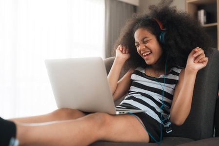 Happy cheerful young black girl using laptop computer on sofa at home enjoy listening the music and singing alongの写真素材