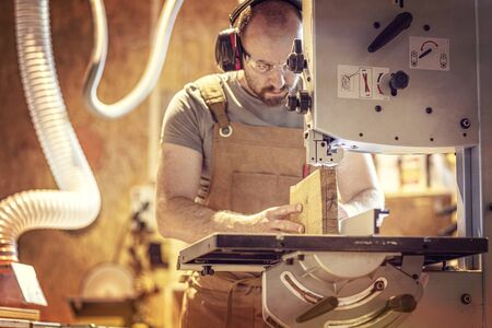 portrait of a carpenter inside his carpentry workshop using a band saw. Wear ear and eye protection