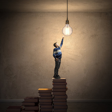boy climbs a ladder of books and reaches for a light bulb, a concept of creativity and educationの写真素材