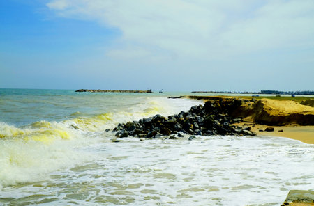 Sea waves cover sand beach and hit black stone pile on shore under blue sky backgroundの写真素材