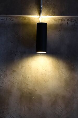 A black cylindrical wall lamp that shines up and down, splashes light onto the bare cement wall of the house decorated in a loft style.の写真素材