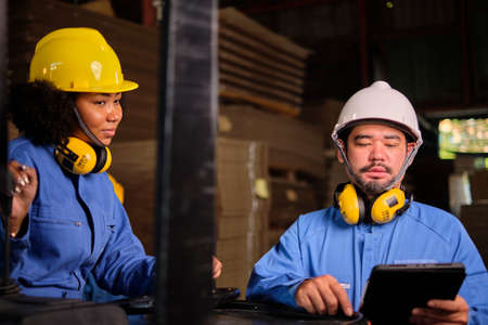 Asian male engineer in safety uniform and hard hat and female worker colleague inspect storage, stock order at factory warehouse, piles of stacking cardboard manufacture, industry product management.の写真素材