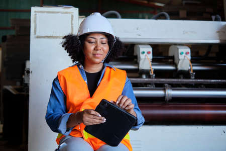African American female engineer worker in safety uniform and hard hat, quality inspect by tablet, maintenance, check the machine at paper production manufacture industry, factory technician manager.の写真素材