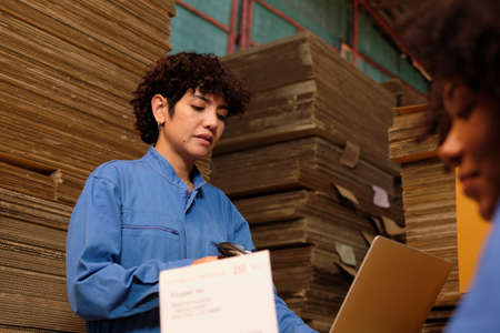 Two safety uniform female workers and colleagues use bar code scanner to check shipment orders stock at parcels warehouse, paper manufacture factory for packing industry, logistic transport service.の写真素材