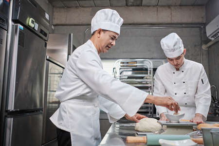 Two professional Asian male chefs in white cook uniforms and aprons are kneading pastry dough and eggs, preparing bread and fresh bakery food, baking in oven at stainless steel kitchen of restaurant.の写真素材