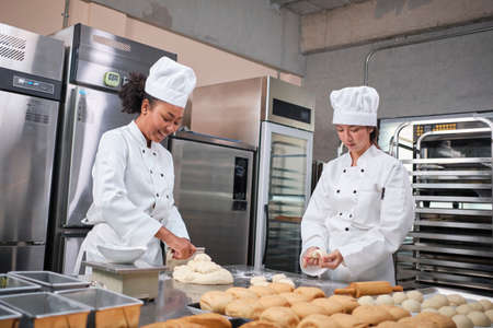 Two professional female chefs in white cook uniforms and aprons knead pastry dough and eggs, prepare bread, cookies, and fresh bakery food, baking in oven at a stainless steel kitchen of a restaurant.の写真素材