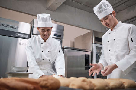 Two professional Asian male chefs in white cook uniforms and aprons are kneading pastry dough and eggs, preparing bread and fresh bakery food, baking in oven at stainless steel kitchen of restaurant.の写真素材