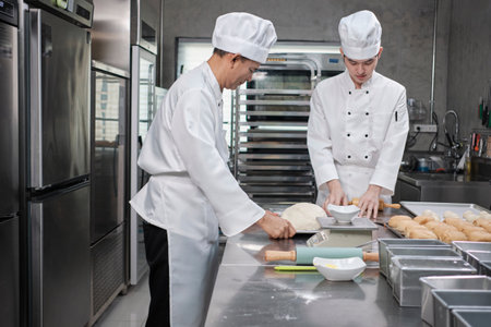 Two professional Asian male chefs in white cook uniforms and aprons are kneading pastry dough and eggs, preparing bread and fresh bakery food, baking in oven at stainless steel kitchen of restaurant.の写真素材