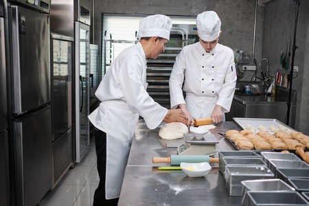 Two professional Asian male chefs in white cook uniforms and aprons are kneading pastry dough and...