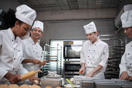 Multiracial professional gourmet team, four chefs in white cook uniforms and aprons knead pastry dough and flour, prepare bread, and bakery food, baking in oven at stainless steel restaurant kitchen.の写真素材