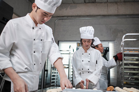Professional gourmet team, three young chefs in white cook uniforms and aprons knead pastry dough and eggs, prepare bread, and fresh bakery food, baking in oven at stainless steel restaurant kitchen.の写真素材