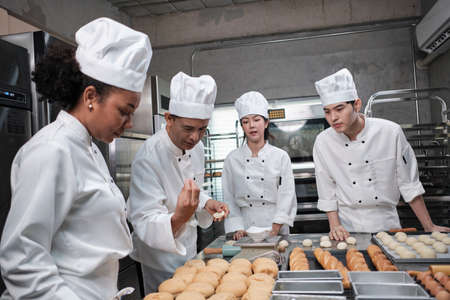 Multiracial professional gourmet team, four chefs in white cook uniforms and aprons knead pastry dough and flour, prepare bread, and bakery food, baking in oven at stainless steel restaurant kitchen.の写真素材