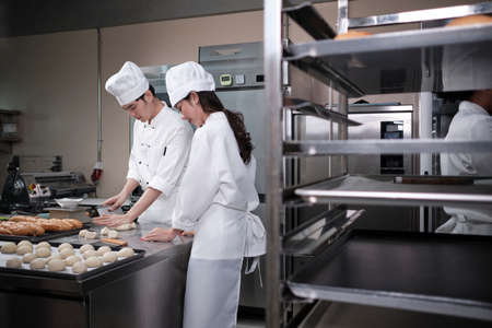 Two professional Asian chefs in white cook uniforms and aprons knead pastry dough and eggs, prepare bread, cookies, and fresh bakery food, baking in an oven in a restaurant stainless steel kitchen.の写真素材