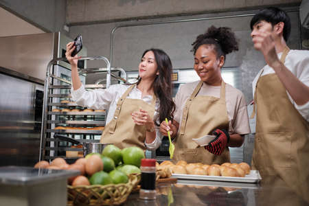 Three young students in cooking class wear aprons enjoyment while taking selfie photo with mobile phone in kitchen, smiling and laughing, preparing eggs and fruits, learn fun culinary course together.の写真素材