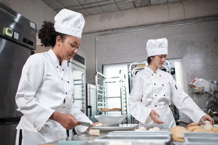 Two professional female chefs in white cook uniforms and aprons knead pastry dough and eggs, prepare bread, cookies, and fresh bakery food, baking in oven at a stainless steel kitchen of a restaurant.の写真素材