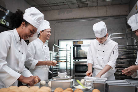 Multiracial professional gourmet team, four chefs in white cook uniforms and aprons knead pastry dough and flour, prepare bread, and bakery food, baking in oven at stainless steel restaurant kitchen.の写真素材