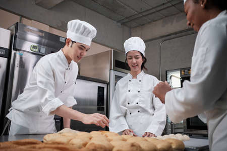 Professional gourmet team, three young chefs in white cook uniforms and aprons knead pastry dough and eggs, prepare bread, and fresh bakery food, baking in oven at stainless steel restaurant kitchen.の写真素材