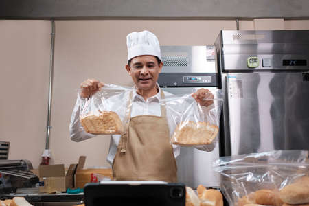 Senior Asian male chef in apron, small business blogger live streaming via mobile application technology, bakery sales online promotion, fresh bread, pastry food, and commercial cooking in kitchen.の写真素材