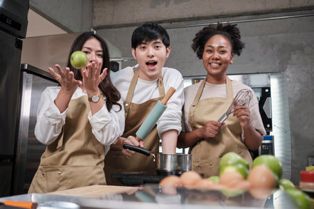Three young students in cooking class wear aprons enjoy and cheerful and fun with ingredients and tools in kitchen, happy smiling, preparing fruits for learning fun culinary course together.の写真素材