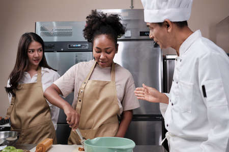 Cuisine course, senior male chef in cook uniform teaches young cooking class students to knead and roll pastry dough, prepare ingredients for bakery foods, fruit pies in stainless steel kitchen.の写真素材