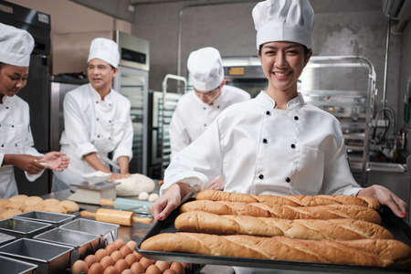 Portrait of young Asian female chef in white cooking uniform looking at camera with a cheerful smile and proud with tray of baguette in kitchen, pastry foods professional and fresh bakery occupation.の写真素材