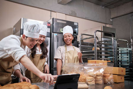 Young three chefs startup partners, small business blogger live streaming via mobile application technology, bakery sales online promotion, fresh bread, pastry food, and commercial kitchen cooking.の写真素材