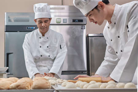Two professional Asian male chefs in white cook uniforms and aprons are kneading pastry dough and eggs, preparing bread and fresh bakery food, baking in oven at stainless steel kitchen of restaurant.の写真素材