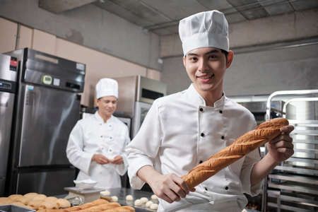 Portrait of young Asian male chef in white cooking uniform looks camera, happy smile and cheerful with baguette, professional foods occupation, commercial pastry culinary jobs in a restaurant kitchen.の写真素材