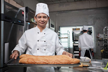Senior Asian male chef in white cook uniform and hat showing tray of fresh tasty bread with a smile, looking at camera, happy with his baked food products, professional job at stainless steel kitchen.の写真素材