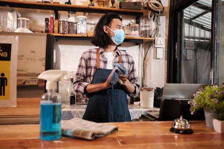 Alcohol sanitizer for healthy protection in a cafe, Asian male barista in face mask use laptop, waiting for coffee orderの写真素材