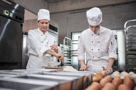 Two professional Asian male chefs in white cook uniforms and aprons are kneading pastry dough and eggs, preparing bread and fresh bakery food, baking in oven at stainless steel kitchen of restaurant.の写真素材