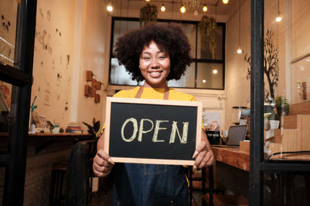 One African American female startup barista stands at casual cafe door, looks at camera, and shows open sign, happy and cheerful smiles with coffee shop service jobs, and new business entrepreneurs.の写真素材