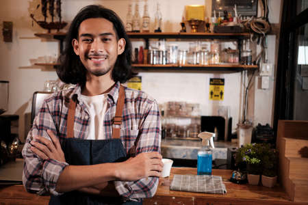 Long hair handsome Asian male startup barista with apron stands at casual cafe counter bar, arms crossed, looks at camera with and welcoming smile, happy and cheerful with coffee shop service jobs.の写真素材