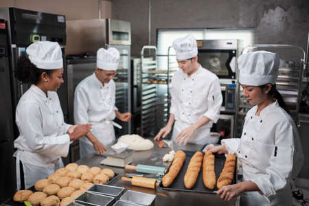 Multiracial professional gourmet team, four chefs in white cook uniforms and aprons knead pastry dough and flour, prepare bread, and bakery food, baking in oven at stainless steel restaurant kitchen.の写真素材