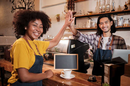 Two cafe business startup partners and friends, African American female, and Thai male baristas look at camera, hand in hand, and cheerful smiles at coffee shop counter bar, happy service staff job.の写真素材