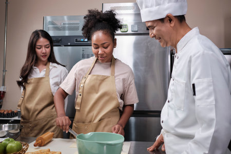 Cuisine course, senior male chef in cook uniform teaches young cooking class students to knead and roll pastry dough, prepare ingredients for bakery foods, fruit pies in stainless steel kitchen.の写真素材