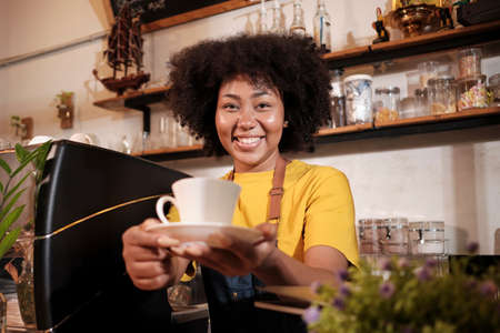 African American female barista in looks at camera, offers cup of coffee to customer with cheerful smile, happy service works in casual restaurant cafe, young small business startup entrepreneur.の写真素材