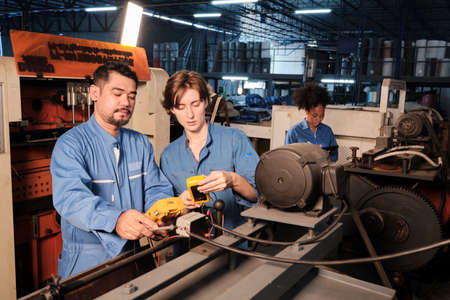 Asian male and White female engineer workers in safety uniforms work by inspecting machines' voltage...