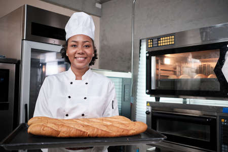 Portrait of African American female chef in white cooking uniform looking at camera with cheerful smile and proud with tray of baguette in kitchen, pastry foods professional, fresh bakery occupation.の写真素材