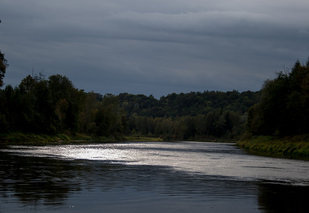 view on river and forests in summer still evening with light reflection in water and cloudy dark sky
