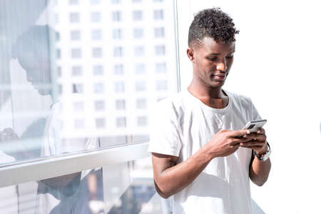 Minimal looking portrait of young African Black man, he is texting a message with a mobile phone in...