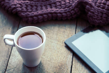 White cup of tea, digital tablet and scarf on old wooden desk. Selective focus, tonedの写真素材