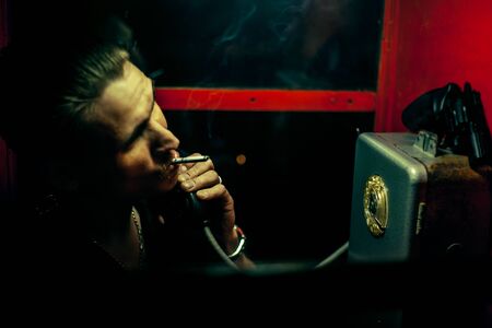 A man of criminal appearance in a payphone booth at night. Talking on the phone with a revolver and...