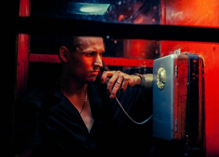 A man of criminal appearance in a payphone booth at night. Talking on the phone with a revolver and...