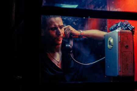 A man of criminal appearance in a payphone booth at night. Talking on the phone with a revolver and a cigarette.の写真素材