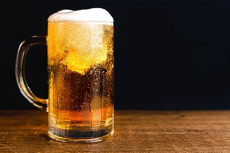 Cold beer with foam in a mug, on a wooden table and a dark background with blank space for text. Stock Photo mug of cold foamy beer close-up.の写真素材