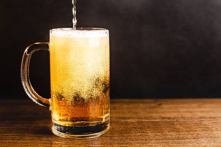 Cold beer with foam in a mug, on a wooden table and a dark background with blank space for  text. Stock Photo mug of cold foamy beer close-up.の写真素材